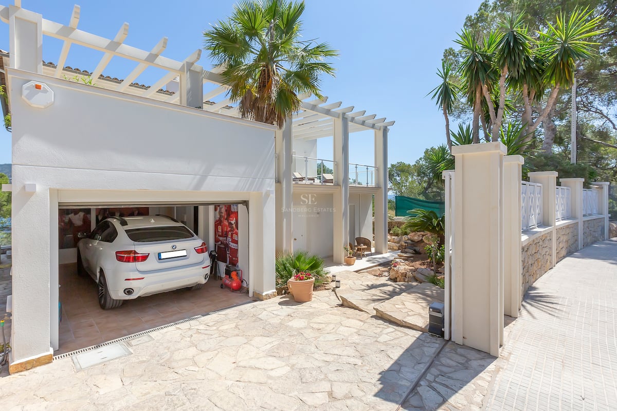 Extérieur d'une villa blanche avec allée en pierre naturelle, pergola et garage ouvert abritant une voiture de luxe.