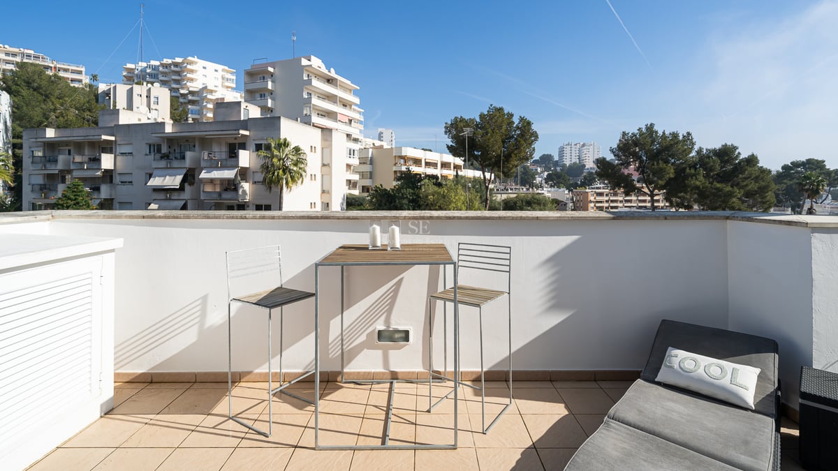 Terrasse ensoleillée avec table haute en bois, deux tabourets et un bain de soleil gris surplombant la ville.