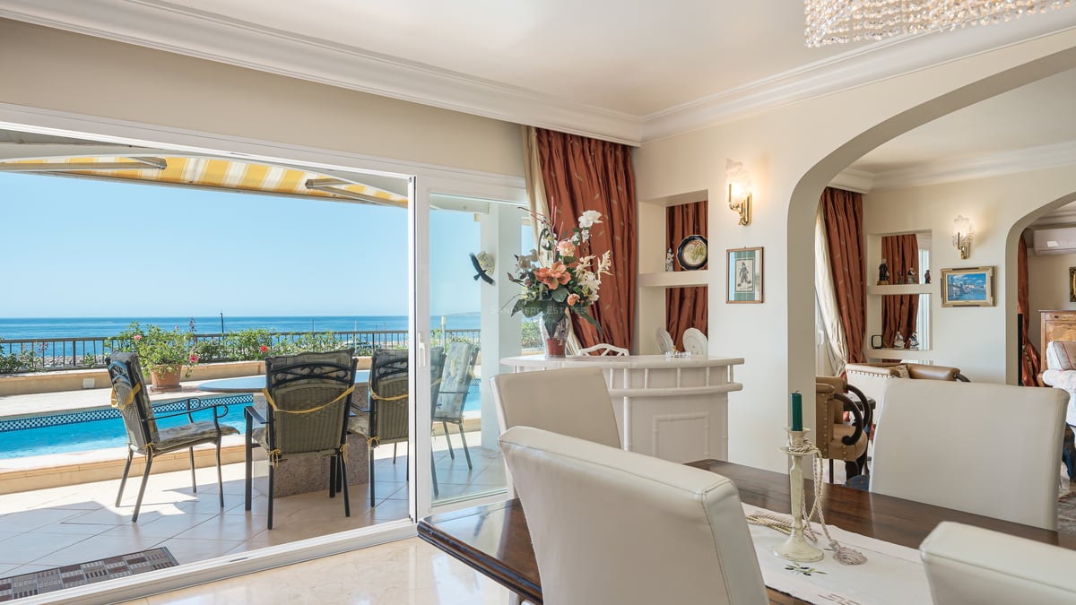 Salle à manger intérieure avec table en bois et chaises blanches donnant sur une terrasse avec piscine et vue mer.