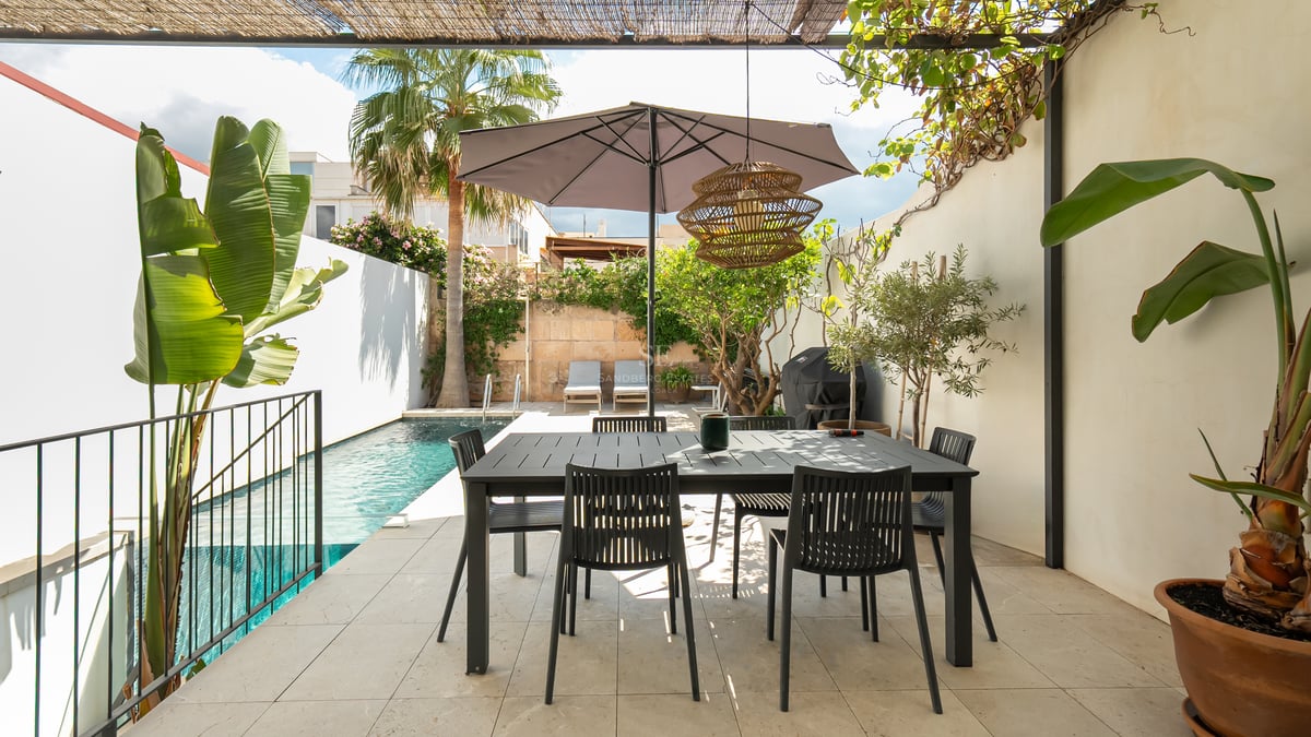 Terrasse moderne avec table à manger noire sous un parasol, à côté d'une piscine turquoise et de palmiers.