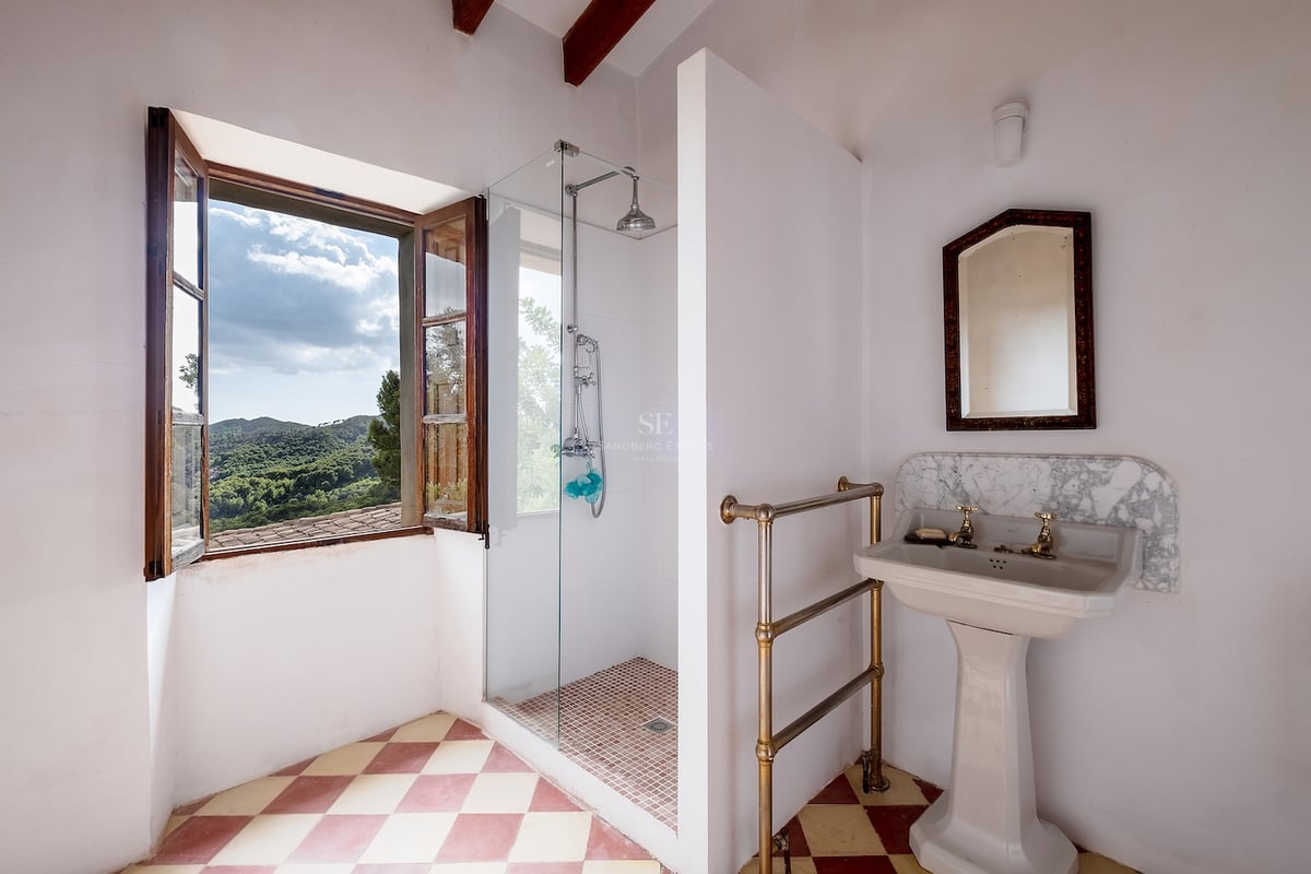 Salle de bain avec sol en damier, lavabo sur pied et douche vitrée près d'une fenêtre donnant sur les montagnes.