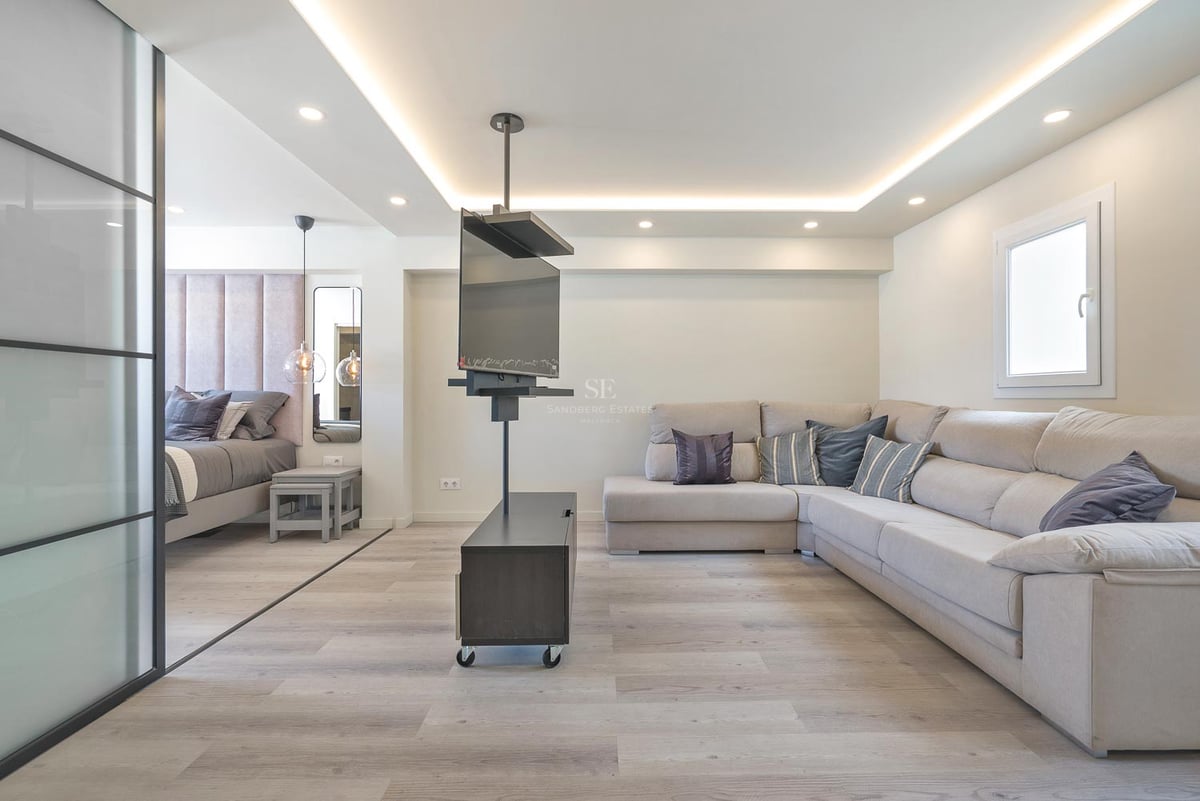 Salon contemporain avec grand canapé en L beige, support TV pivotant et chambre adjacente séparée par une paroi vitrée.