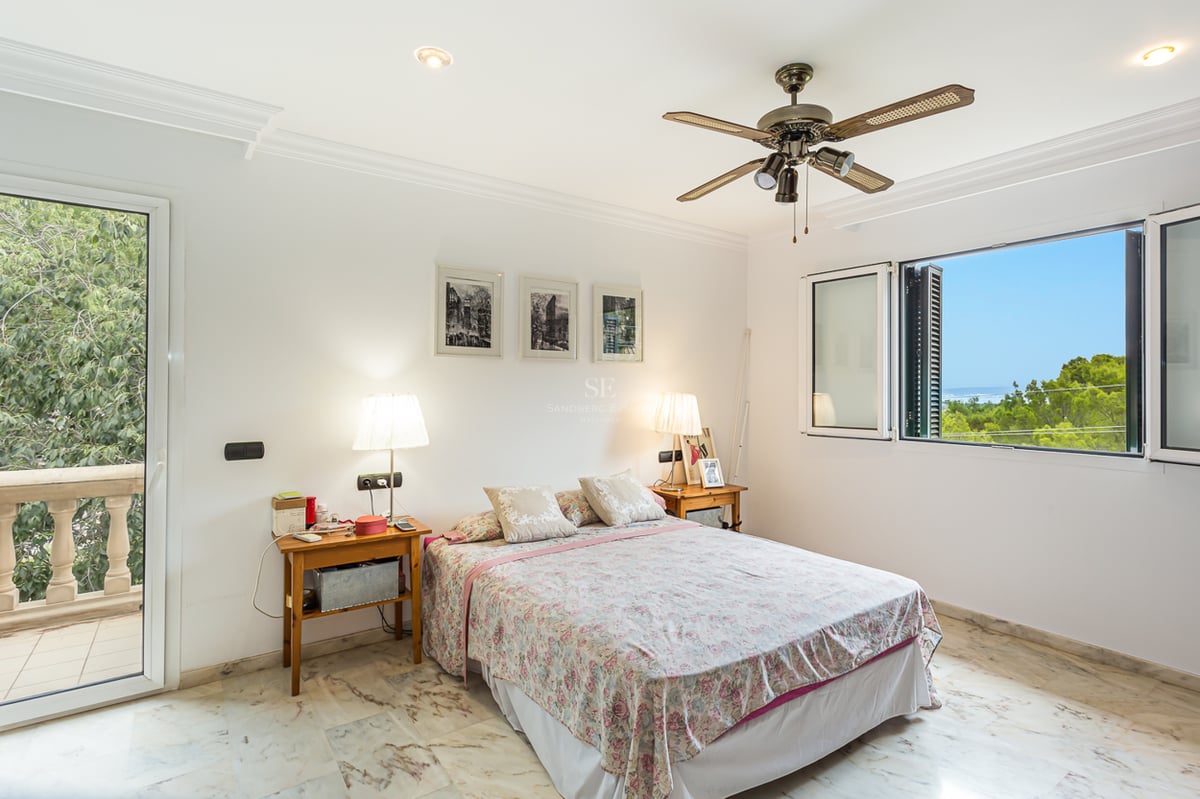 Chambre blanche avec sol en marbre, lit double, ventilateur de plafond et fenêtre offrant une vue sur la mer.