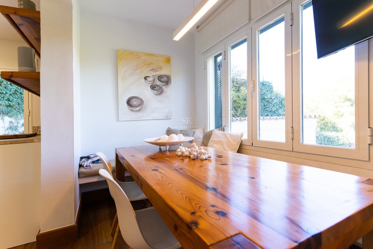 Coin repas avec une grande table en bois, chaises blanches et banquette avec coussins près d'une fenêtre ensoleillée.