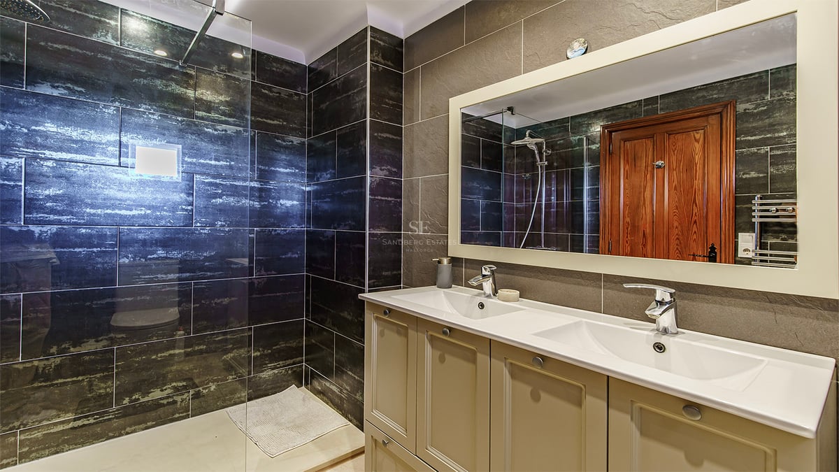 Salle de bains moderne avec double vasque, grand miroir et douche vitrée avec carrelage mural sombre texturé.