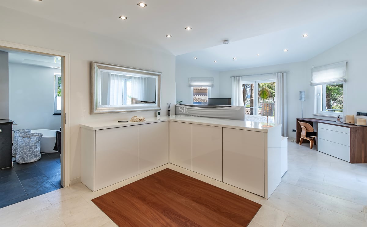 Grande chambre moderne avec rangements blancs, grand miroir, sol en pierre et vue sur la salle de bains attenante.