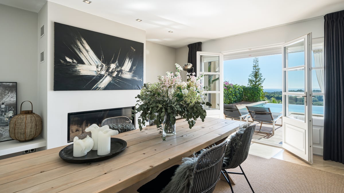 Salle à manger avec table en bois et cheminée ouvrant sur une terrasse avec piscine à débordement et vue mer.