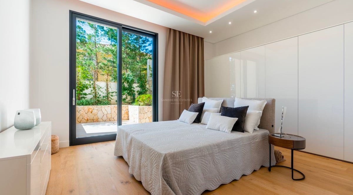 Chambre lumineuse avec grand lit, parquet en bois et baie vitrée coulissante donnant sur un patio privé verdoyant.