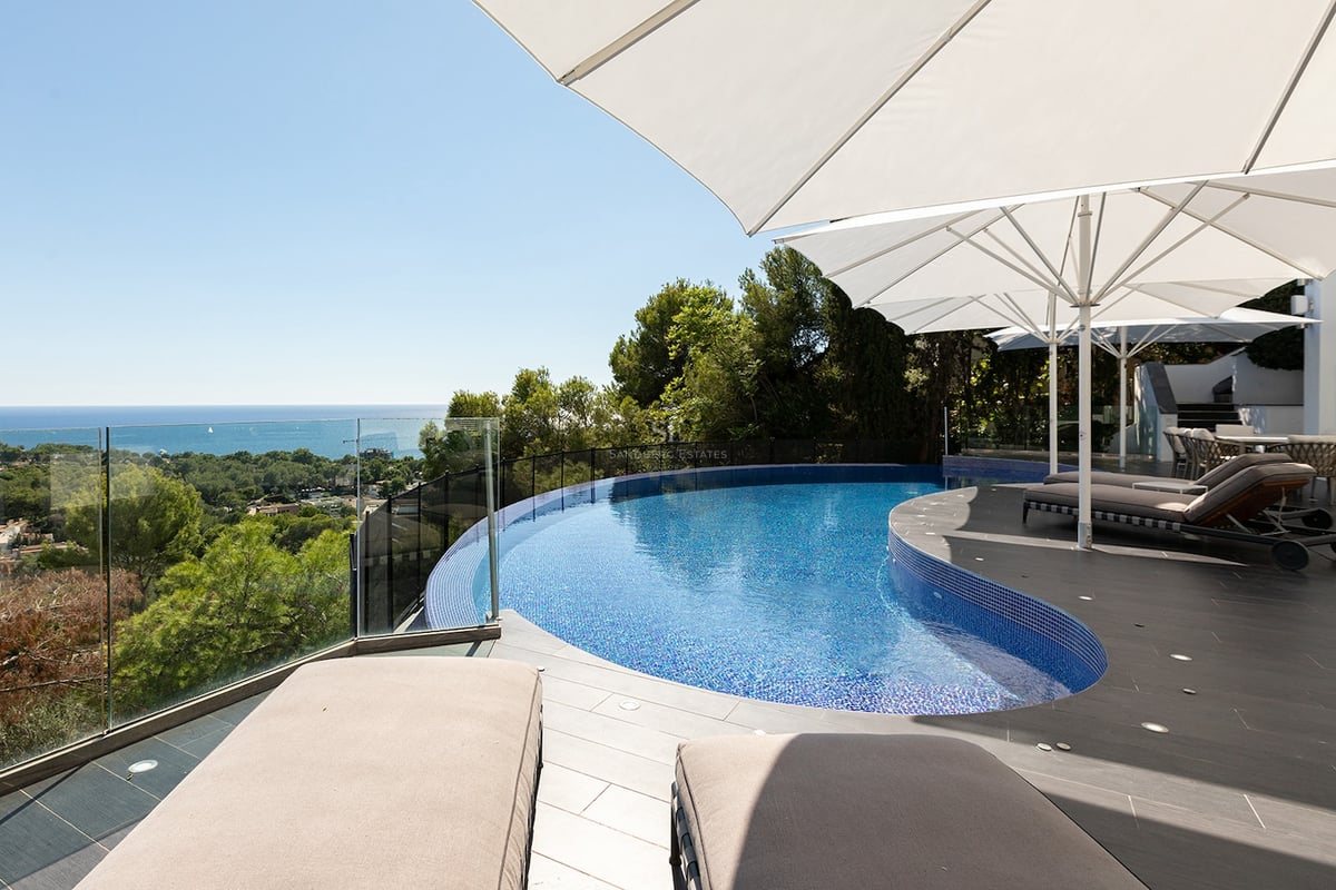 Piscine bleue incurvée sur une terrasse en pierre avec parasols blancs et transats face à la mer.
