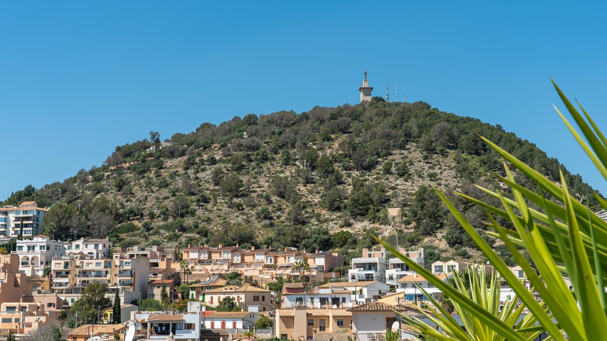 Vue de bâtiments résidentiels méditerranéens devant une colline verdoyante avec un monument sous un ciel bleu clair.