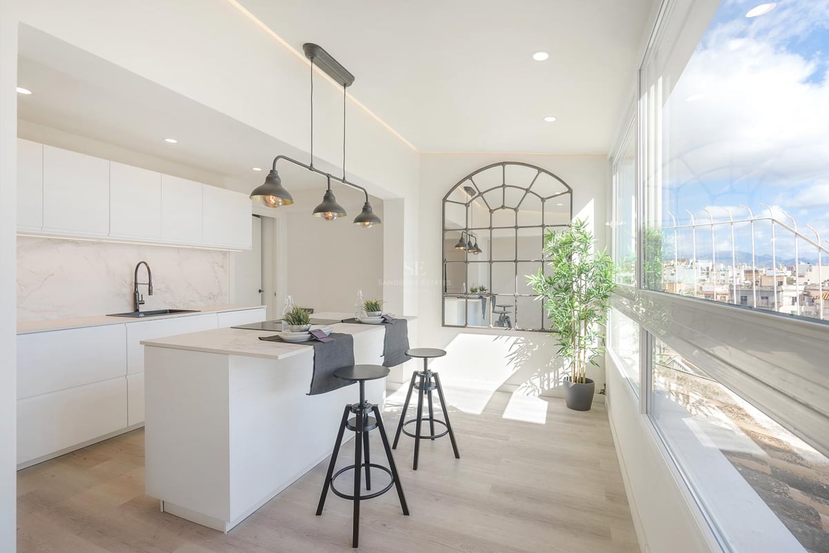 Cuisine moderne lumineuse avec îlot blanc, tabourets industriels noirs et grandes fenêtres donnant sur la ville.