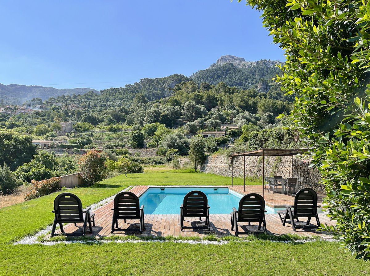 Piscine rectangulaire avec terrasse en bois et transats face aux montagnes verdoyantes sous un ciel bleu azur.