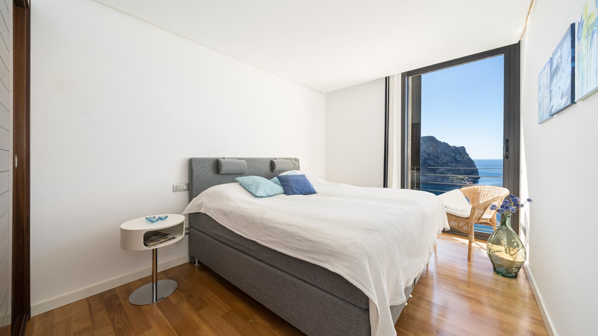 Chambre contemporaine avec lit gris, parquet en bois et baie vitrée offrant une vue sur la mer et les falaises.