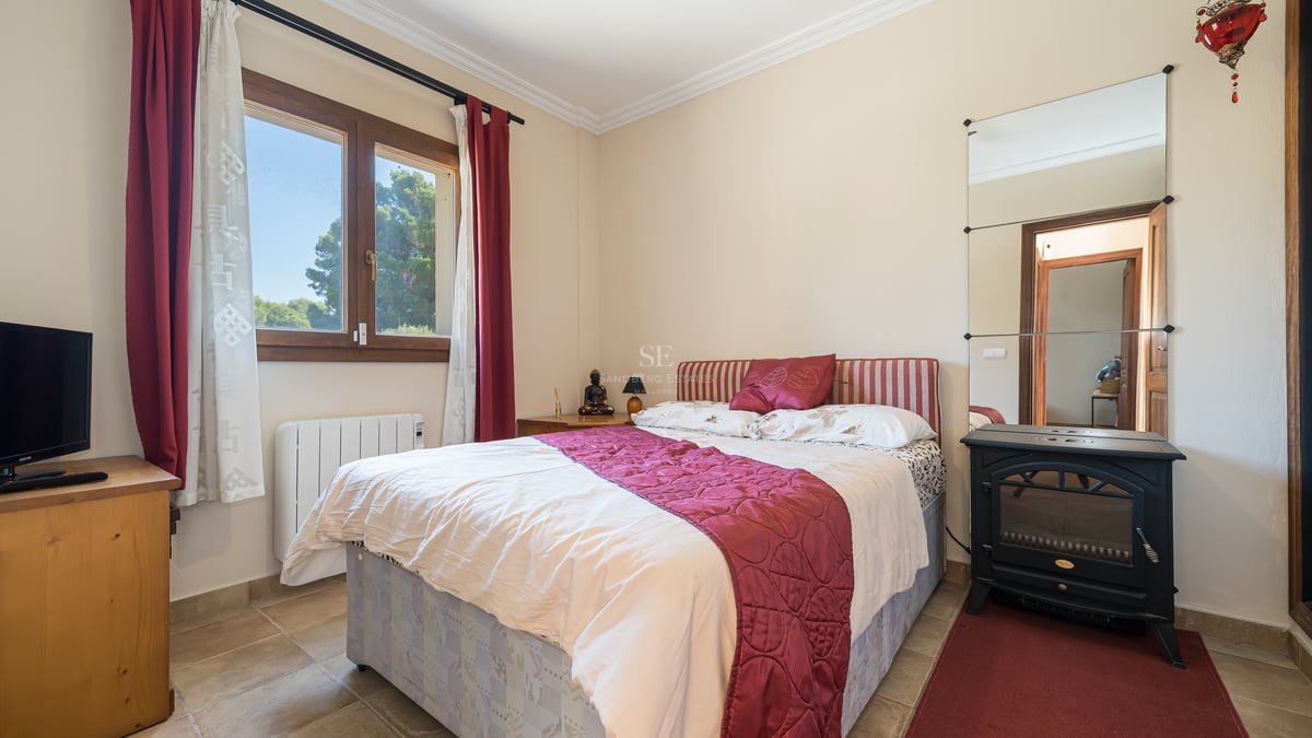 Chambre avec lit double, literie bordeaux, fenêtre en bois vue sur les pins et poêle noir décoratif.