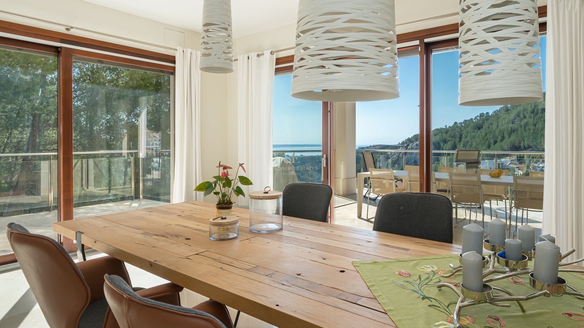Salle à manger moderne avec table en bois rustique, chaises en cuir et baies vitrées donnant sur la côte.