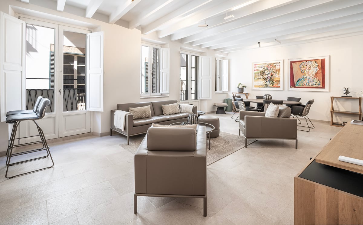 Grand salon avec canapés en cuir taupe, plafond à poutres blanches et grandes fenêtres avec volets.