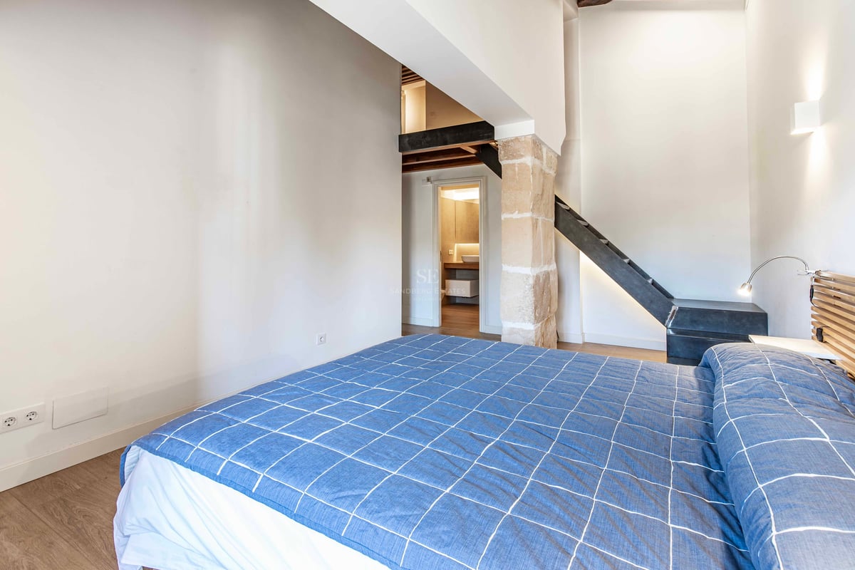 Chambre moderne avec lit bleu, pilier en pierre apparente et escalier menant à une mezzanine.