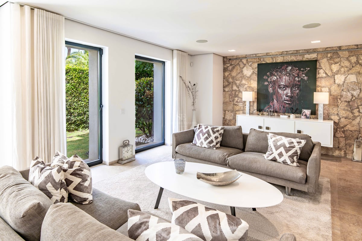 Salon lumineux avec canapés gris, mur d'accent en pierre et baies vitrées donnant sur le jardin.