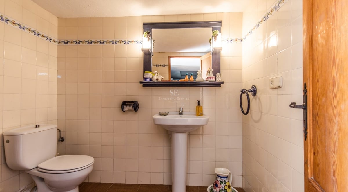 Salle de bain carrelée en crème avec lavabo sur colonne, toilettes et miroir à cadre sombre avec éclairage intégré.