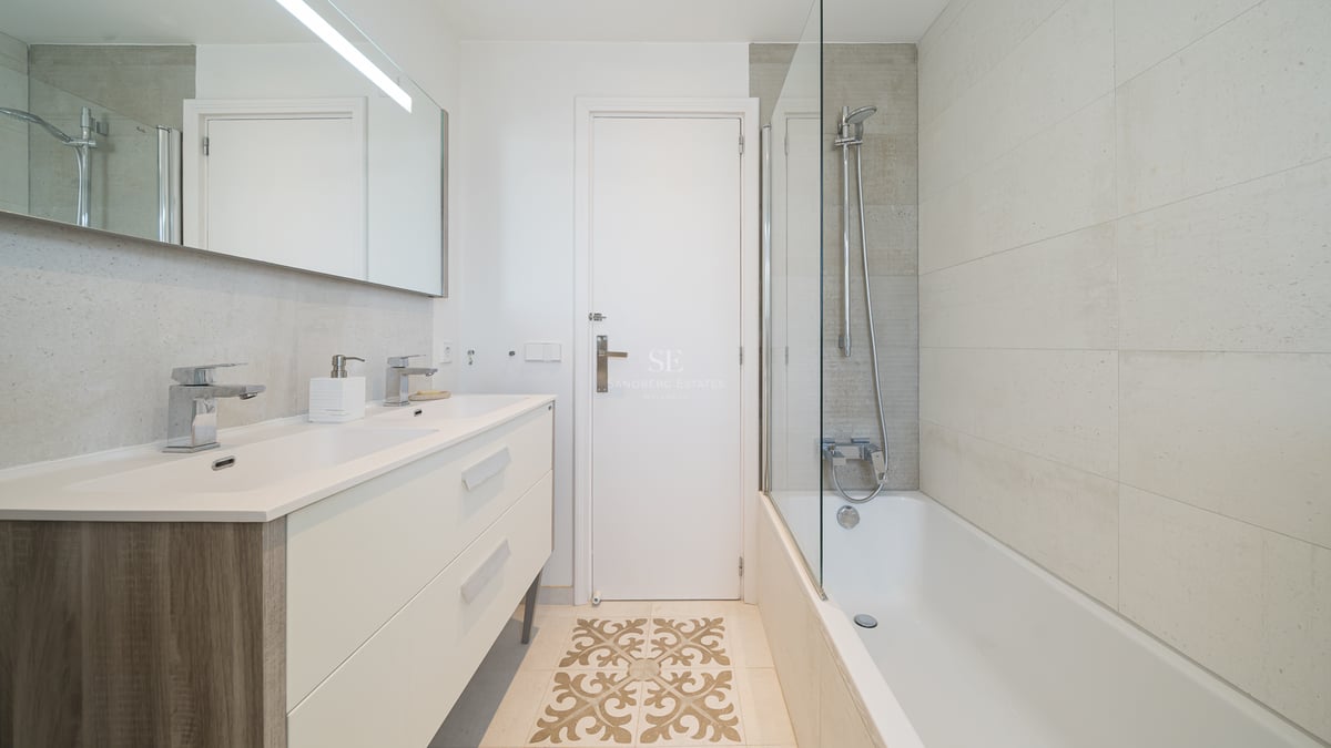 Salle de bain moderne avec double vanité crème, baignoire avec paroi vitrée et carrelage décoratif au sol.