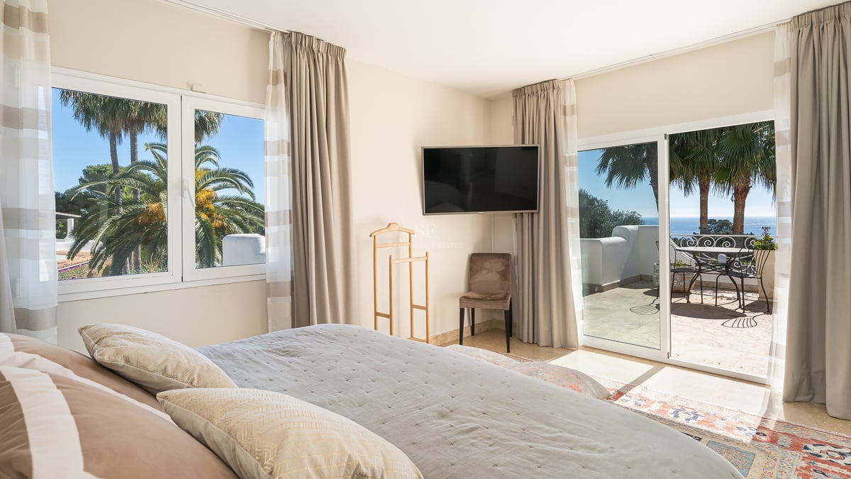 Chambre principale moderne avec grand lit, TV murale et baies vitrées ouvrant sur une terrasse ensoleillée vue mer.