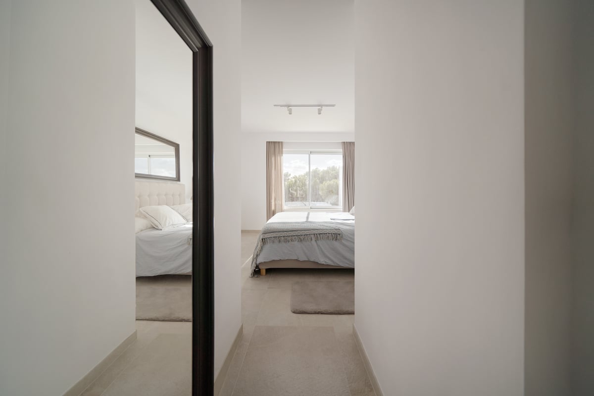 Vue d'un couloir vers une chambre ensoleillée avec grande fenêtre, parure de lit sobre et miroir mural.