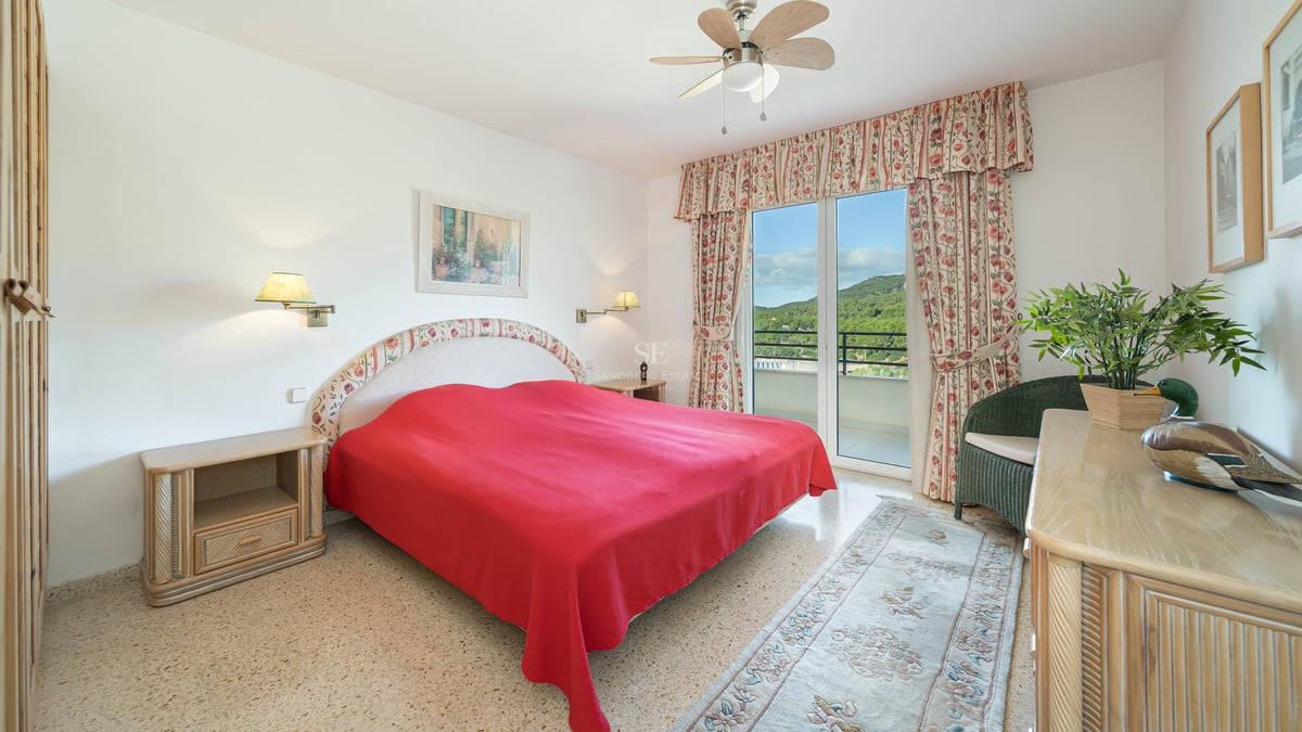 Chambre lumineuse avec couvre-lit rouge, rideaux floraux et balcon offrant une vue sur la montagne.