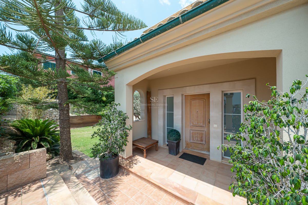 Entrée de villa voûtée avec porte en bois, sol en terre cuite et végétation luxuriante sous un ciel dégagé.