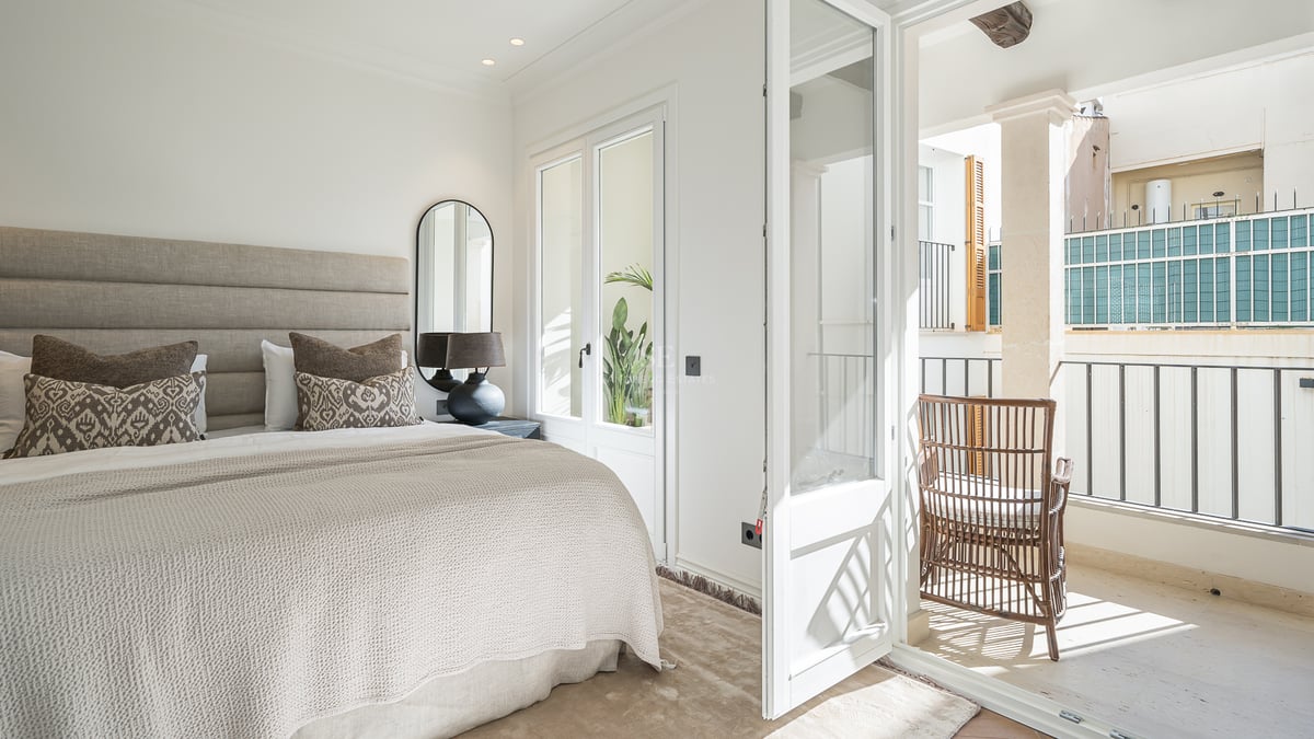 Bright bedroom with neutral linens, upholstered headboard, and open glass doors leading to a stone-tiled balcony.