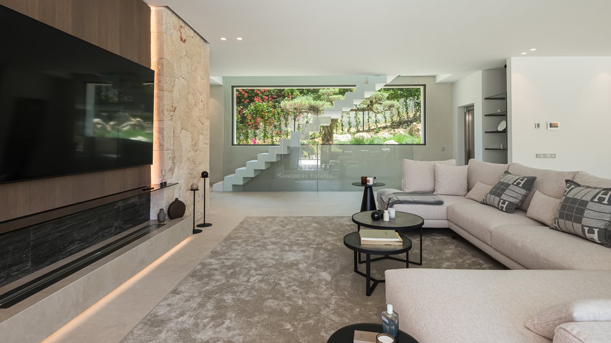 Salon moderne avec grand canapé, mur en pierre naturelle, TV intégrée et escalier suspendu en verre.