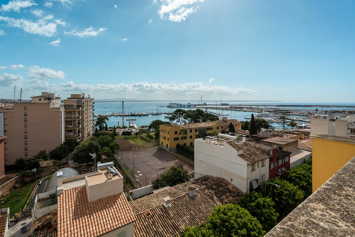 Vue surélevée d'un port méditerranéen avec des yachts, des toits en terre cuite et la mer bleue sous un ciel clair.