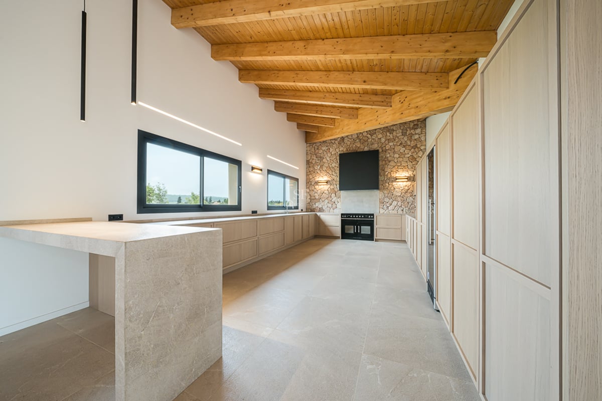 Cuisine moderne spacieuse avec hauts plafonds boisés, mur en pierre naturelle, armoires beiges et électroménager noir.