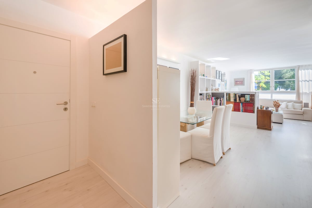 Une entrée blanche et lumineuse menant à un salon minimaliste avec du parquet en bois clair et de grandes fenêtres.