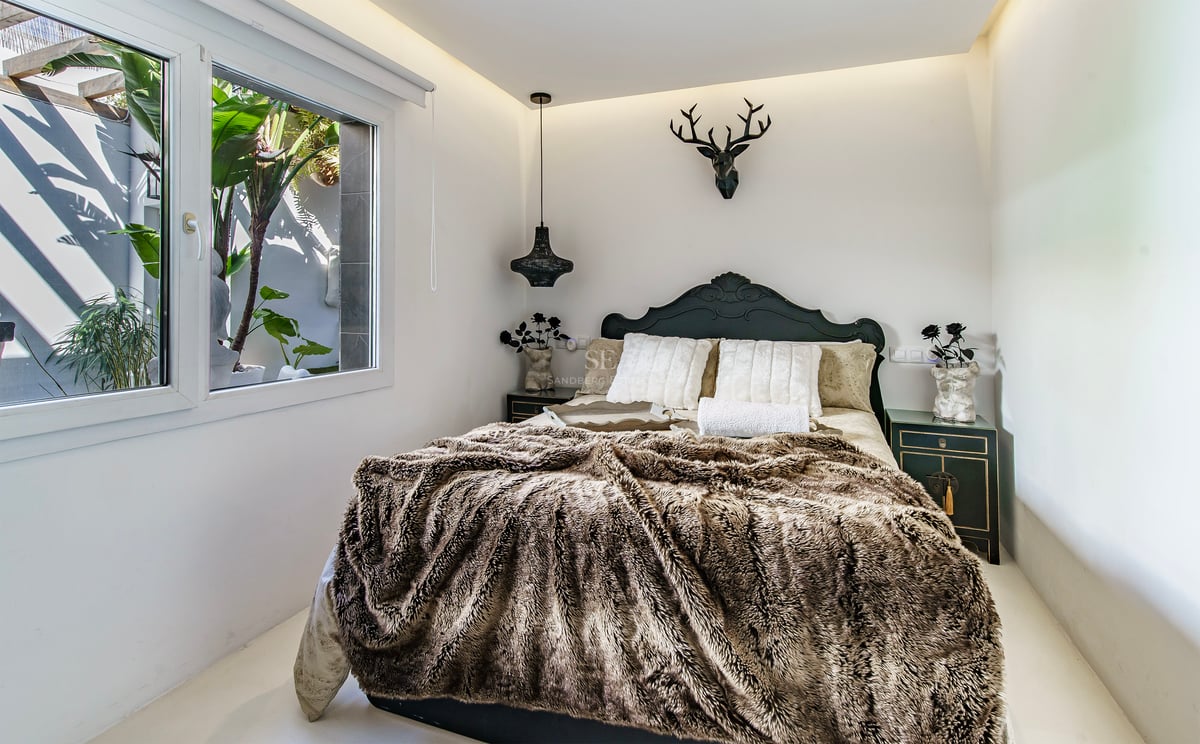 Chambre avec jeté de lit en fourrure, tête de lit sombre, tête de cerf décorative et fenêtre sur verdure.