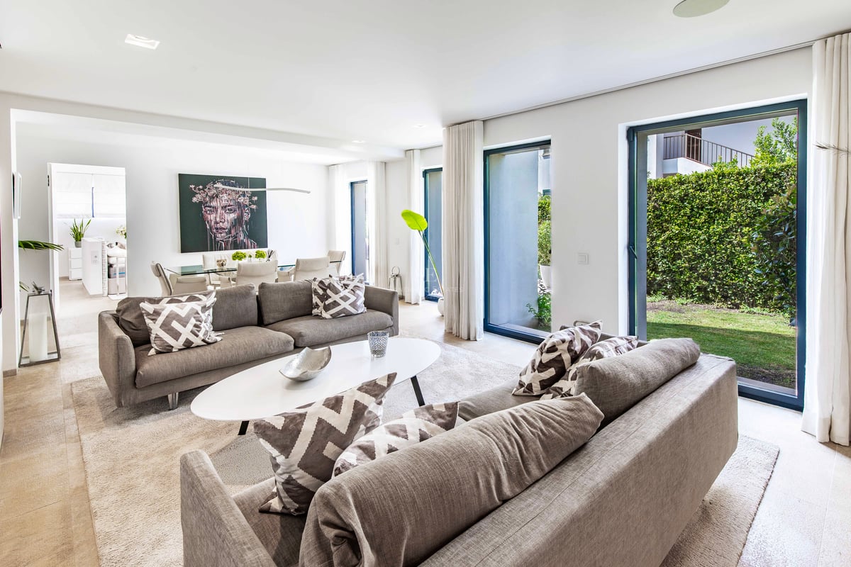 Salon lumineux avec deux canapés gris, table basse blanche et grandes baies vitrées ouvrant sur un jardin privé.
