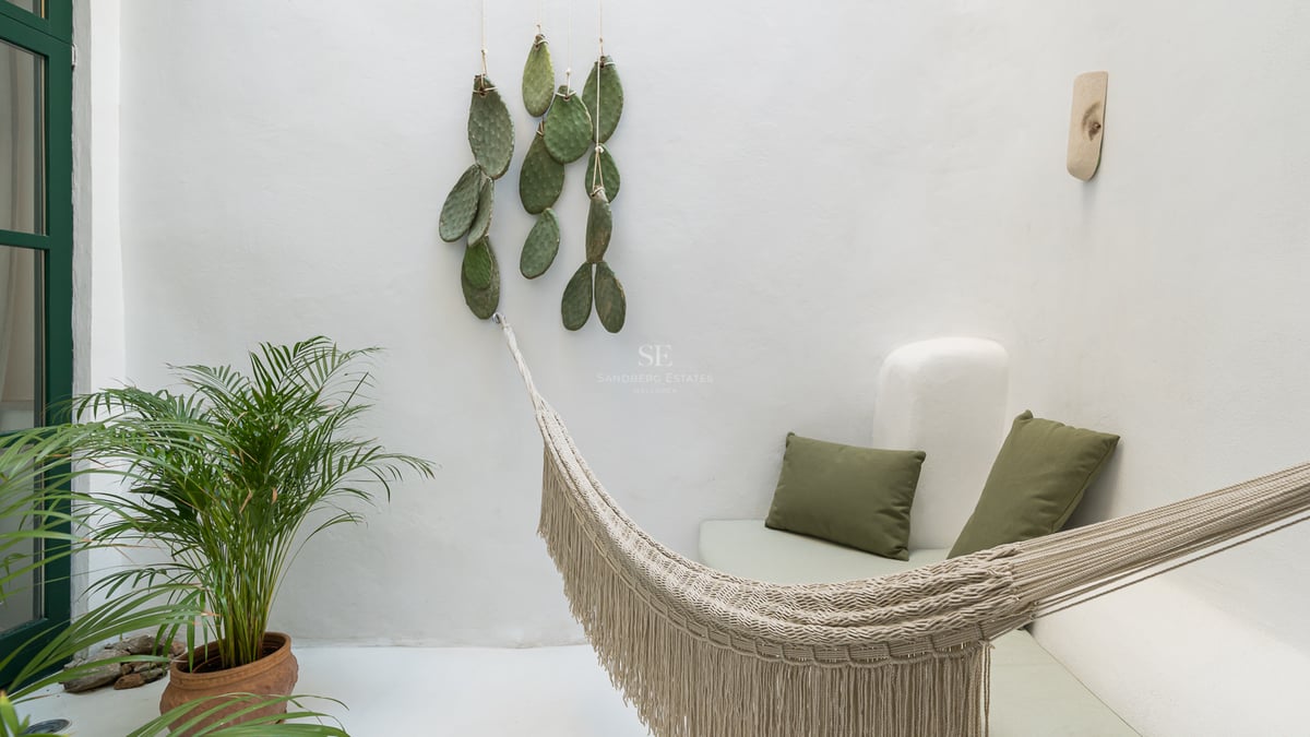 Un hamac tissé crème dans une cour minimaliste blanche avec des coussins verts et des plantes.
