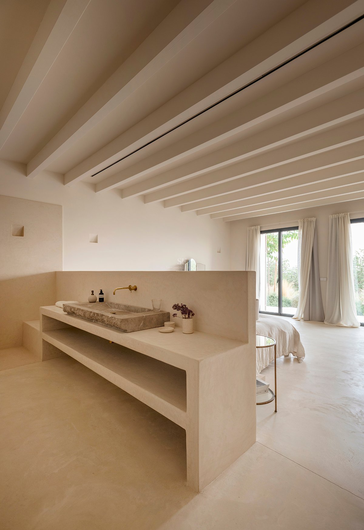 Minimalist bedroom with a built-in stone washbasin, exposed ceiling beams, and floor-to-ceiling glass doors.