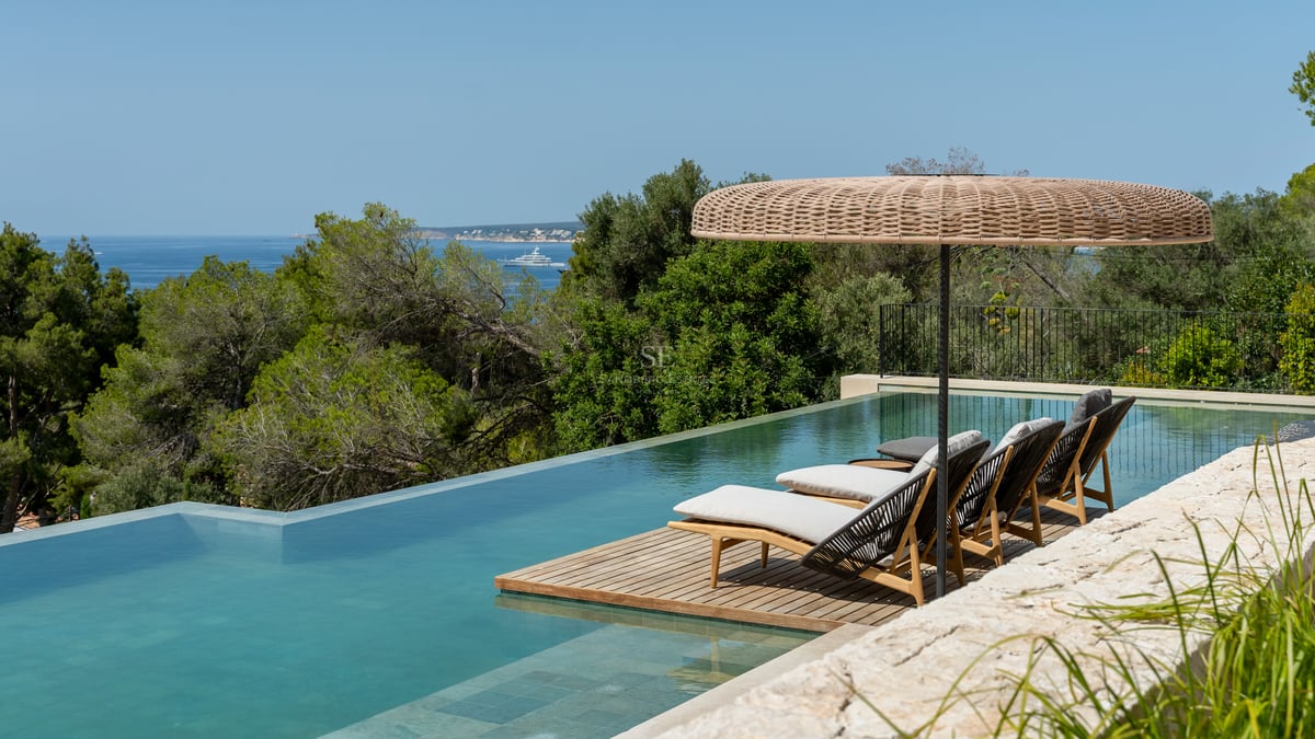 Piscine à débordement avec terrasse en bois, chaises longues et parasol en osier face à la mer et aux arbres.