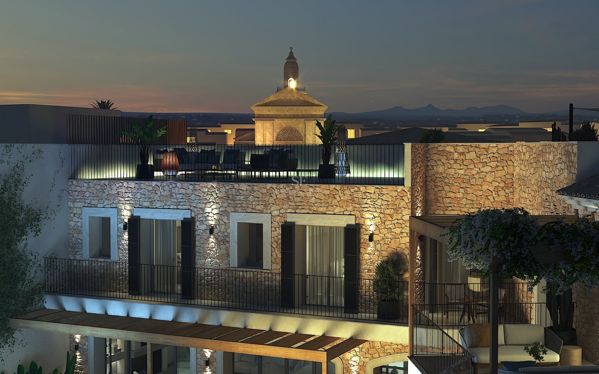 Vista nocturna de una villa mediterránea con paredes de piedra iluminadas y terraza en la azotea con vistas al pueblo.