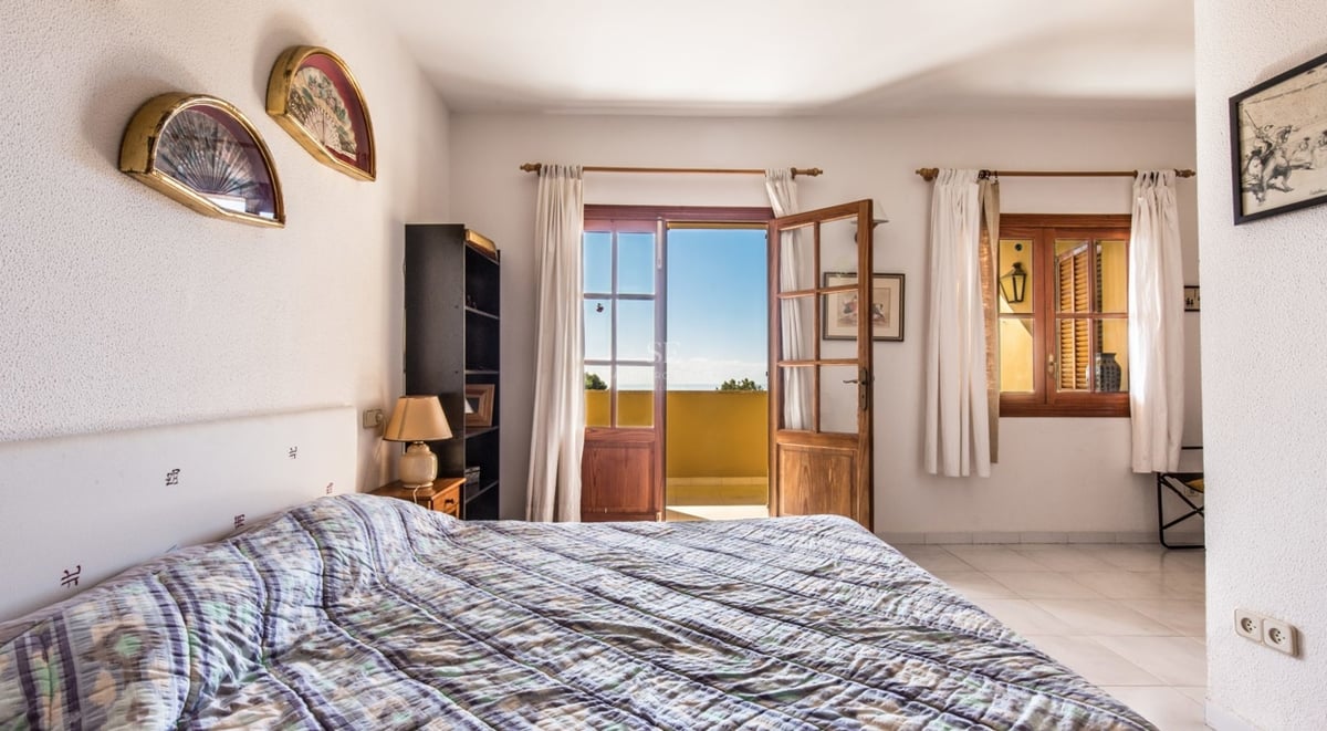 Une chambre lumineuse avec un lit double, des murs blancs texturés et des portes en bois s'ouvrant sur un balcon ensoleillé.