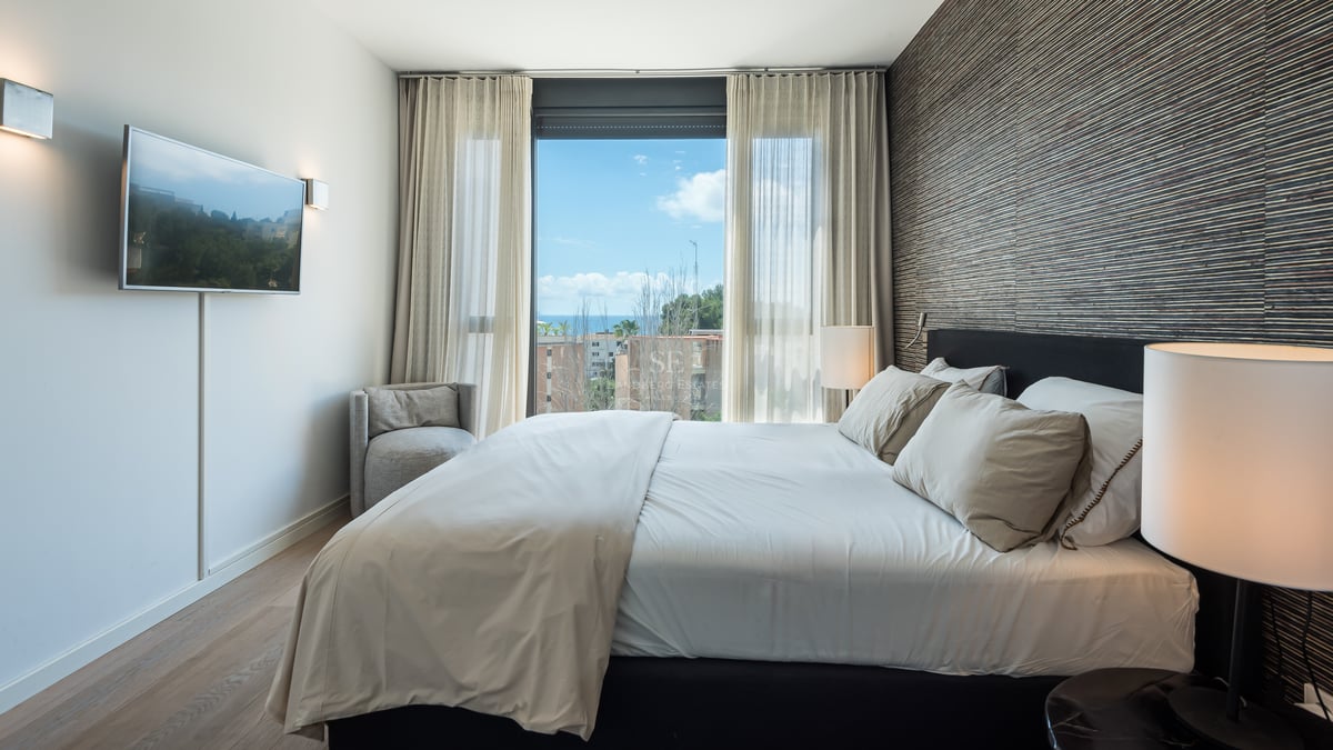 Chambre contemporaine avec grand lit, mur d'accent texturé et fenêtre donnant sur la mer et le ciel bleu.