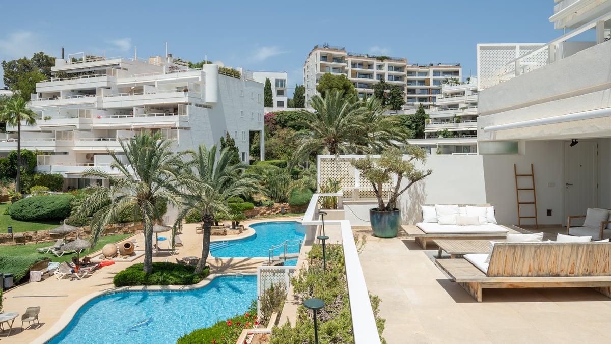 Terrasse ensoleillée avec mobilier en bois surplombant des piscines bleues et des bâtiments blancs méditerranéens.