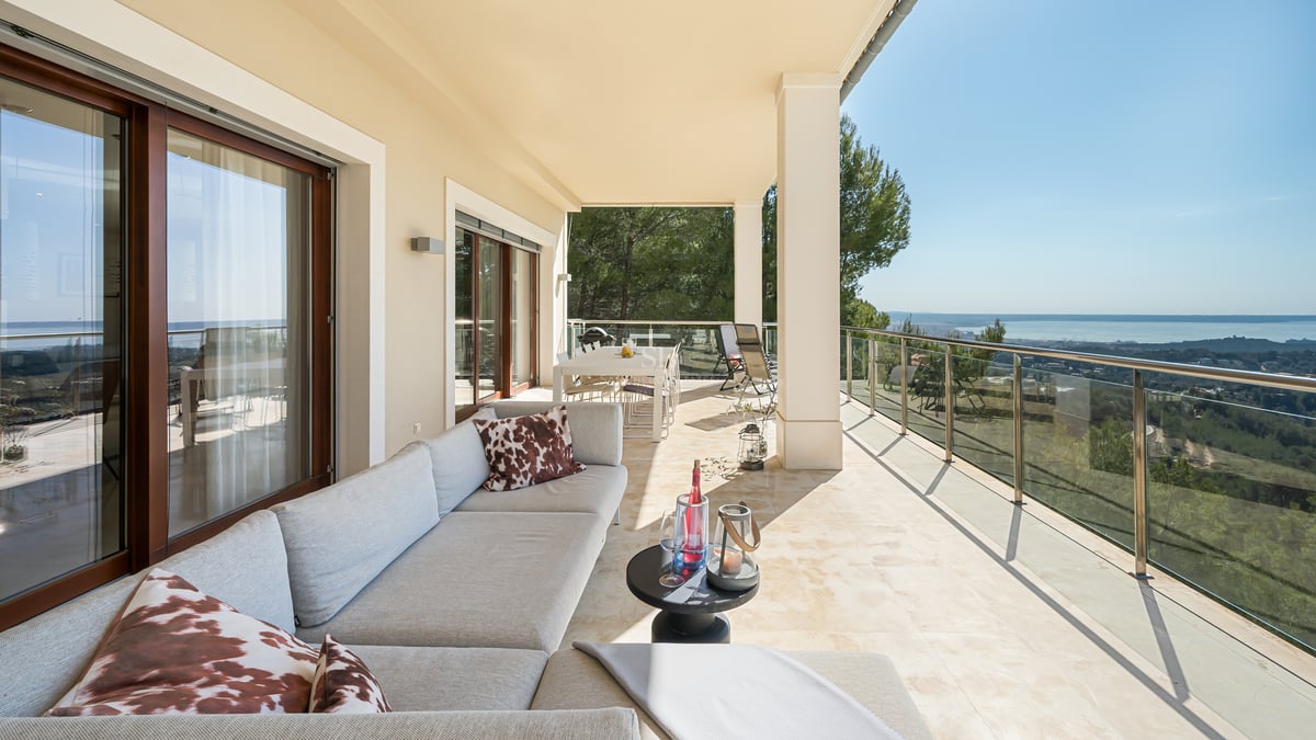 Terrasse de luxe couverte avec sol en pierre naturelle, canapé gris, garde-corps en verre et vue sur la mer et la forêt.