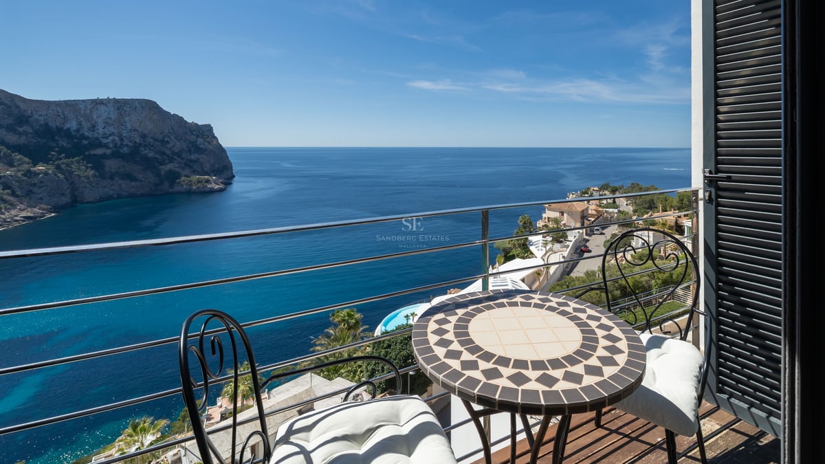 Une petite terrasse avec table en mosaïque et chaises en fer donnant sur une baie méditerranéenne bleue et des falaises.