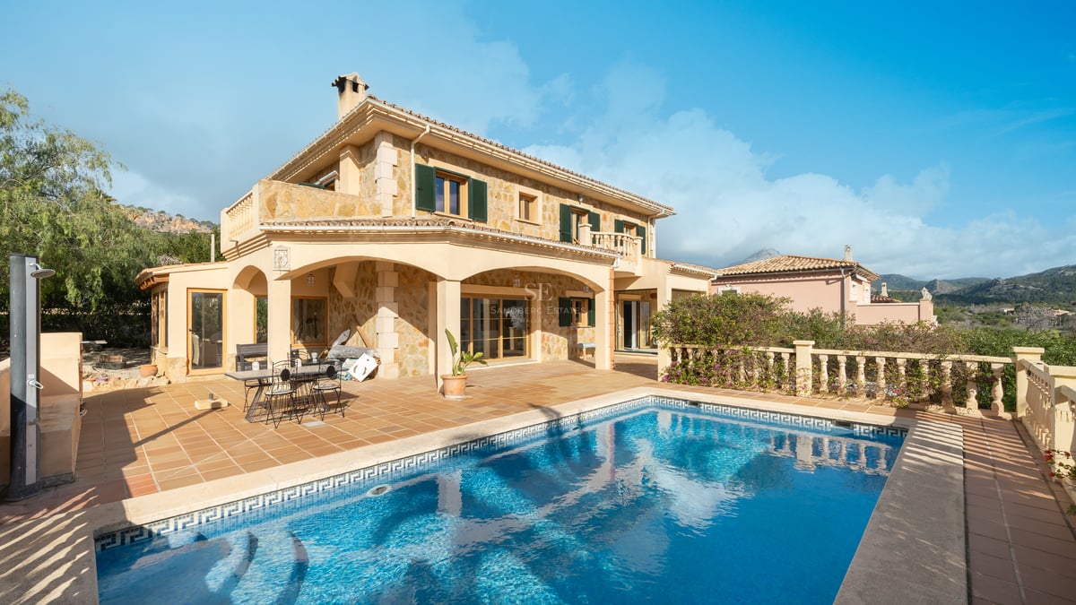 Vue d'une villa en pierre de deux étages avec piscine turquoise et terrasse en terre cuite sous un ciel bleu.