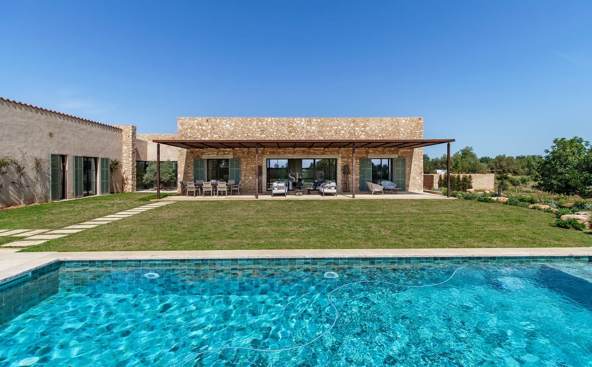 Exterior de villa de lujo con piscina turquesa, césped y fachada de piedra natural bajo un cielo despejado.