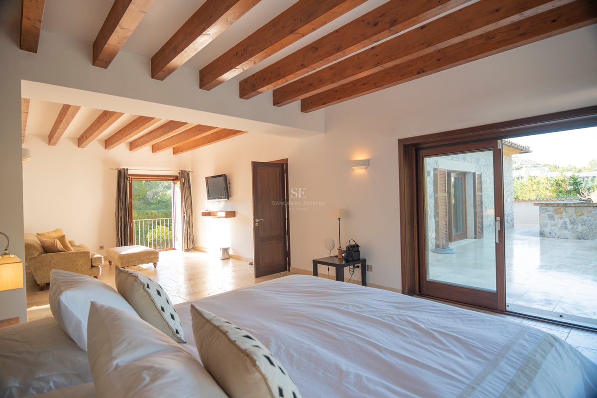 Chambre spacieuse avec hauts plafonds, poutres en bois, sol en pierre et baies vitrées coulissantes menant à la terrasse.