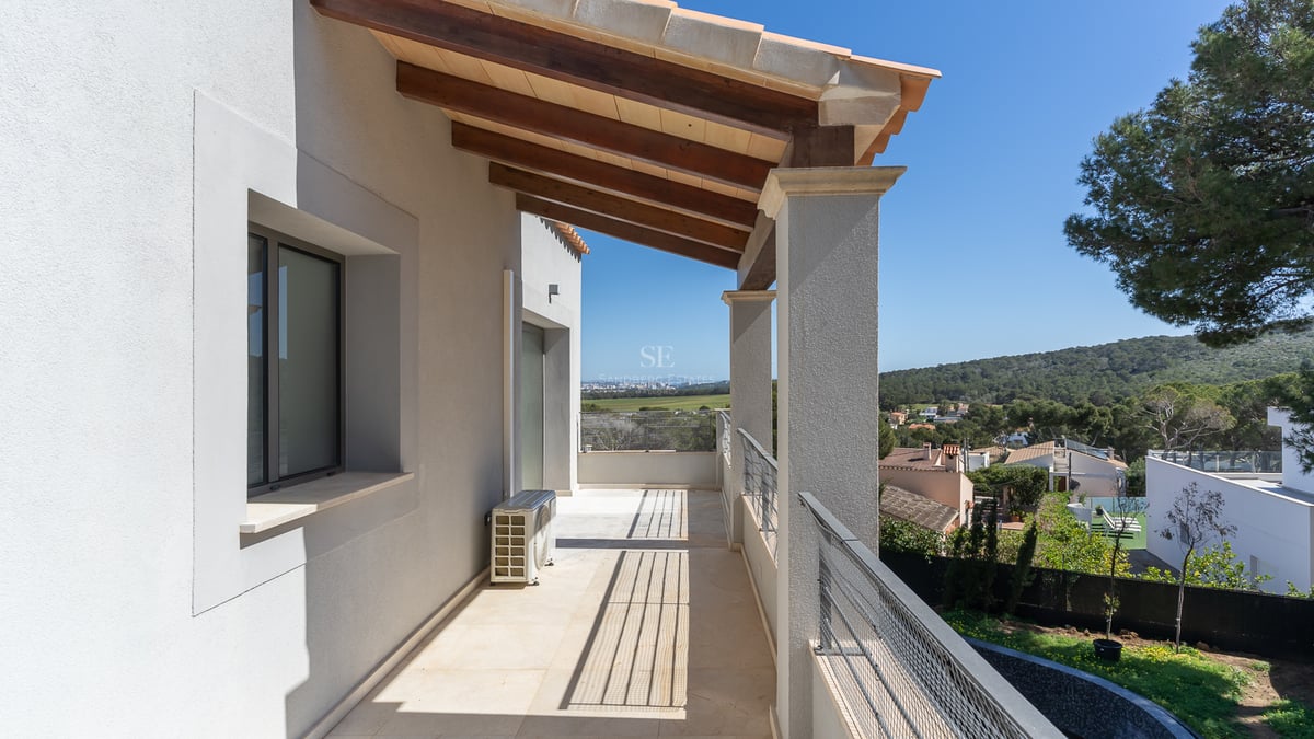 Long balcon couvert avec poutres en bois, murs blancs et garde-corps métallique surplombant une vallée verdoyante.