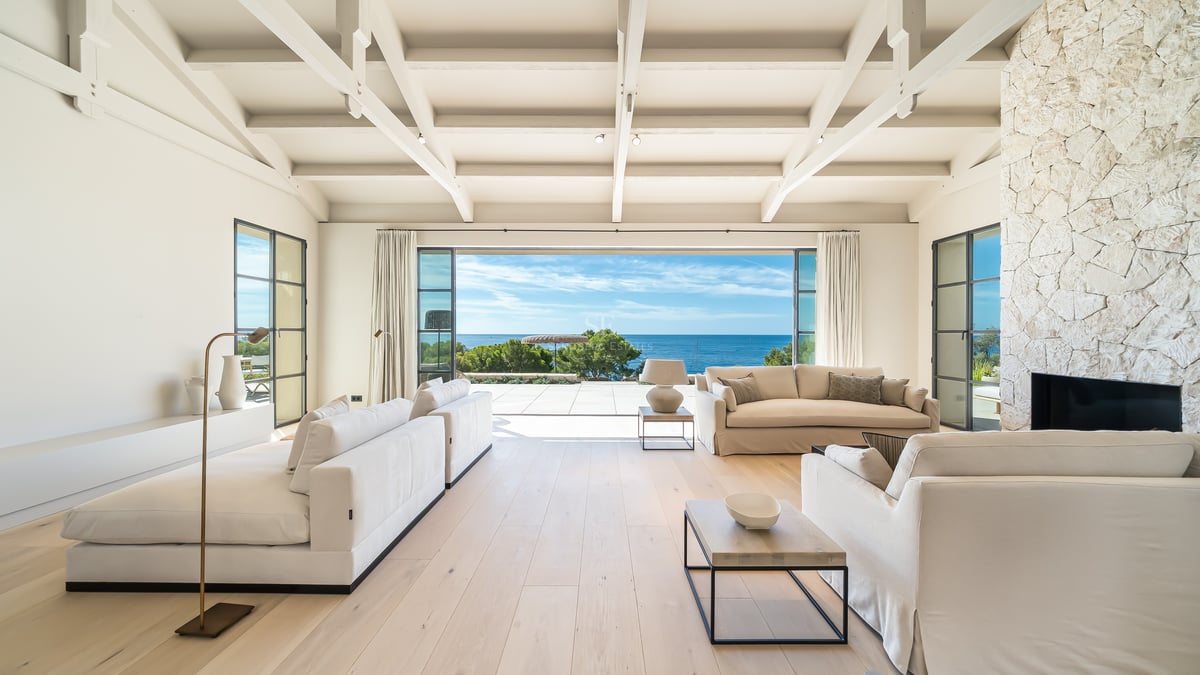 Salon spacieux avec plafonds à poutres blanches, parquet clair et grandes baies vitrées s'ouvrant sur la mer.
