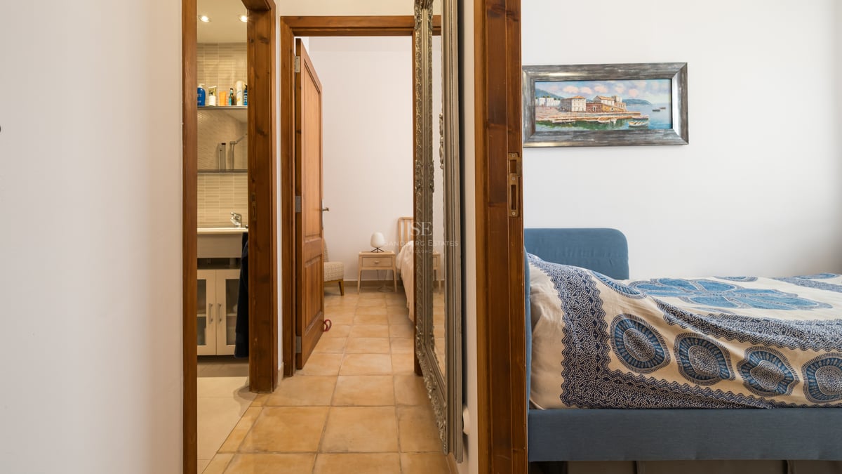 Un couloir avec carrelage en terre cuite menant à une salle de bain et deux chambres, doté d'un grand miroir argenté.